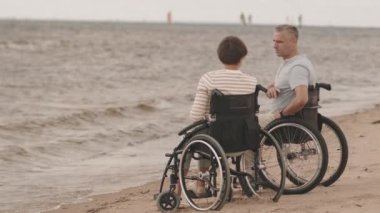 Romantik yetişkin beyaz tekerlekli sandalyeli çift kumsalda deniz kenarında oturmuş gibi konuşuyorlar.