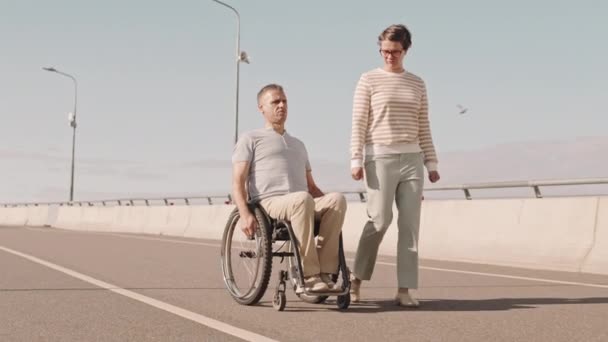 成熟的白人男子坐轮椅沿着桥路行走 而他微笑的妻子则站在他的身边 在户外共度夏天 与他交谈 — 图库视频影像