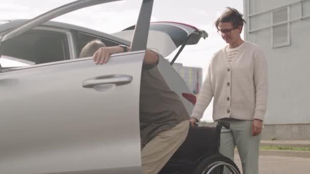 車の運転席から出るために障害のある大人の男性を助ける一方 車椅子を保持する介護女性は外に駐車 — ストック動画