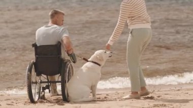 50 'li yaşlarda, kumlu sahilde tekerlekli sandalyede oturan beyaz saçlı bir adam yaz gününü şefkatli karısı ve sevimli açık sarı Labrador av köpeğiyle birlikte geçiriyor.