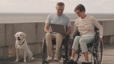 Modern tekerlekli sandalyedeki çift dışarıda dizüstü bilgisayarda sohbet ederken açık sarı Labrador Retriever 'ları yakınlarda dinleniyor.