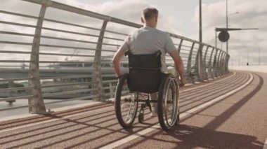 Yazın köprüde koşu pisti boyunca tekerlekli sandalyeye mahkum, olgun, beyaz bir adamın arka görüntüsü.