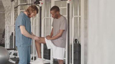 Mavi üniformalı profesyonel fizyoterapi uzmanı, ağır yaralı siyah adamın motor sistemini ve bacak gücünü inceliyor. Spor antrenmanına hazırlanıyor.