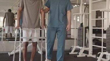 Zombileri ve mavi önlüklü fizyoterapisti olan Afrikalı Amerikalı yavaş yavaş fizyoterapi seansı sırasında spor salonunda birlikte yürüyorlar.
