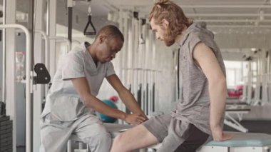 Modern rehabilitasyon merkezinde kişisel fizyoterapi seansı yapan beyaz erkek hasta ile konuşan Afrikalı Amerikalı fizyoterapist.