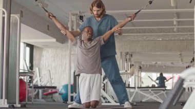 Profesyonel erkek kineziyoterapisti engelli genç siyahi bir adama özel ekipman kullanarak rehabilitasyon egzersizleri yapmasına yardım ediyor.