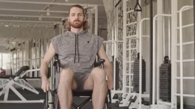 Spor kıyafetli genç beyaz adamın portresi tekerlekli sandalyede oturuyor ve modern fizyoterapi merkezinde rehabilitasyon sırasında kameraya bakıyor.