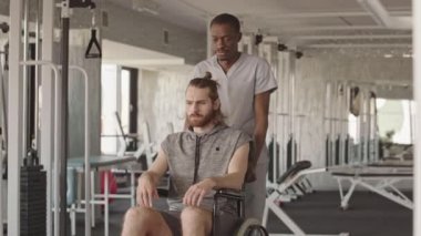 İyimser Afrikalı Amerikalı fizyoterapistin portresi, modern fizyoterapi merkezinde tekerlekli sandalyedeki genç beyaz erkek hastayı taşıyor, kameraya bakıyor ve gülümsüyor.