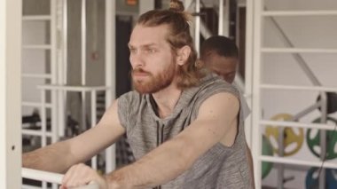 Fizyoterapi merkezinde fizyoterapist ile kişisel seans yaparken sakallı, fizyoterapi simülatörüyle bacak egzersizi yapan beyaz bir adam.