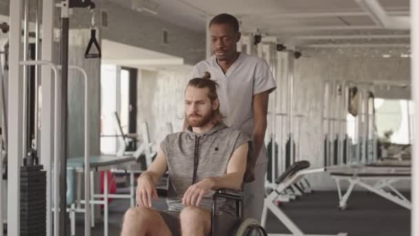 楽観的なアフリカ系アメリカ人理学療法士の肖像現代の理学療法センターで車椅子で若い白人男性患者を運ぶ カメラと笑顔の両方を見て — ストック動画