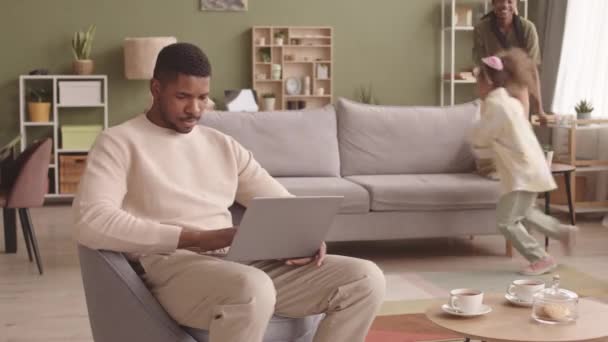 年轻的黑人男子在家里的笔记本电脑上工作 而他的妻子和两个爱玩的小女儿在一起玩 时间过得真慢 — 图库视频影像