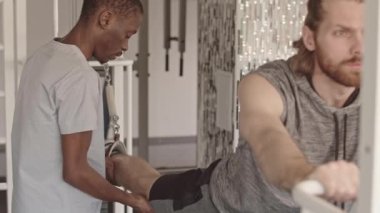 Afrikalı Amerikalı fizyoterapist, rehabilitasyon merkezindeki özel fizyoterapi simülatörü üzerinde bacak egzersizi yapmak için yaralı genç beyaz bir adama yardım ediyor.
