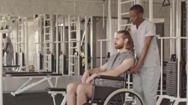 Tekerlekli sandalyedeki genç beyaz adam farklı makine ve ekipmanlarla spor rehabilitasyon merkezinde fizyoterapi koçuyla kişisel seans yapıyor.