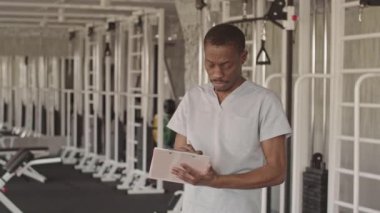 Formalı genç Afrikalı Amerikalı fizyoterapistin orta boy portresi. Kameraya bakıyor, modern rehabilitasyon merkezinde çalışıyor.
