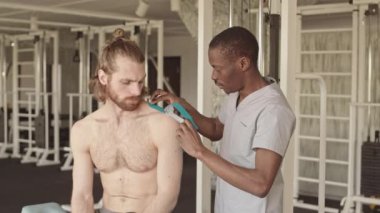 Ameliyat önlüğü takmış Afrikalı Amerikalı fizyoterapistin acıdan omuzlarına mavi kinesio bandı yapıştırması, rehabilitasyon merkezinde spor salonu ile kişisel seans yapması.