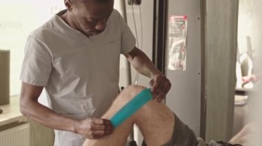 Önlüklü Afrikalı Amerikalı kayropraktik uzmanı, fizyoterapi seansı sırasında tanınmayan hastaların diz eklemine mavi kinesio bandı yapıştırıyor.