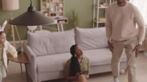一个年轻的黑人男子在客厅里挂着灯 一边慢慢换灯泡 一边和美丽的妻子和两个小女儿在沙发上跑来跑去 玩接球游戏 — 图库视频影像