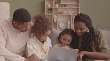 Afro-Amerikan evli çiftin orta yavaş portresi ve kameraya gülümseyen iki küçük kızı kanepede oturmuş kızların kucağında dizüstü bilgisayarlarıyla