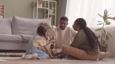 Dört çocuklu mutlu Afro-Amerikan aile oturma odasında oturup el oyunu oynarken başparmaklarını birbirlerine uzatıp yüksek kule yapıyorlar.