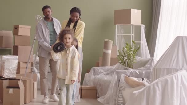 两个漂亮的非洲裔美国小女孩慢吞吞地帮助她们的父母搬着铺满纸板箱的新公寓里的地毯沿着客厅走着 — 图库视频影像
