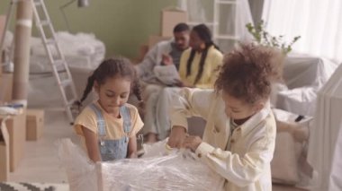 İki güzel Afrikalı Amerikalı kızın yeni daireye taşındıktan sonra naylonları açması ve arka plandaki koltukta ailelerinin sohbet etmesi.