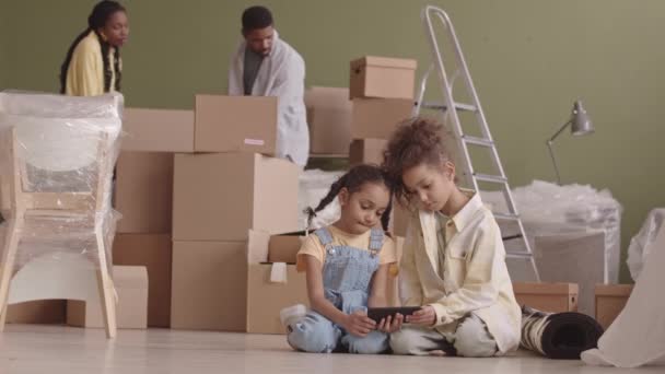結婚したアフリカ系アメリカ人のカップルのスローモは段ボール箱を持って新しいアパートに入り 2人の小さな娘はスマートフォンでビデオを見ながら家具なしのリビングルームの床に座っています — ストック動画
