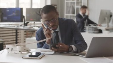 Mavi takım elbiseli Afro-Amerikan erkek borsacıların yavaşlaması akıllı telefon ve dizüstü bilgisayarla online borsayı izlerken telefonda yatırımcılarla konuşuyor, iş arkadaşlarıyla aynı masada oturuyor.