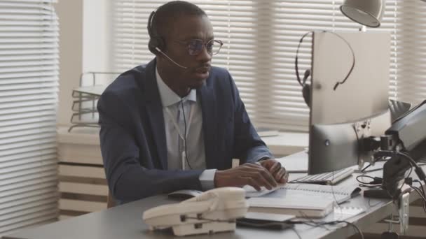 忙碌的非洲裔美国股票经纪人身穿雅致的蓝色西装 头戴耳机 看着电脑显示器 分析加密货币市场的指数 与客户交谈 在办公室工作 — 图库视频影像