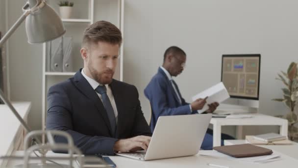 在与非裔美国人商业伙伴一起工作的过程中 穿着西装的英俊白人商人在笔记本电脑上工作 用智能手机打电话的速度有所减慢 — 图库视频影像