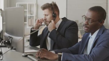 İki farklı borsa simsarı ofiste bilgisayar başında oturmuş borsayı analiz ediyor ve müşterilerle konuşuyor.
