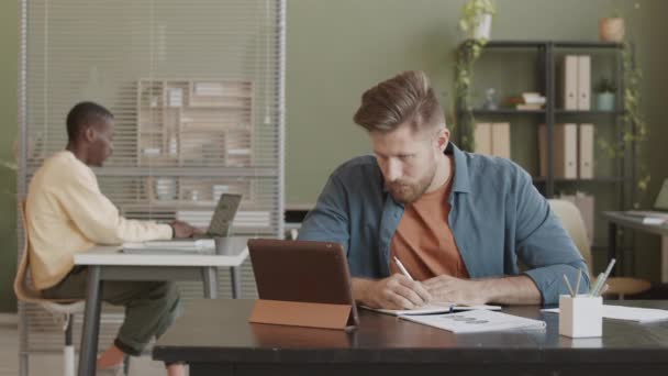 中等速度慢的年轻白人商人在办公室的平板电脑上工作时 随随便便把笔记写在笔记本上 他的非裔美国同事在笔记本电脑上打字的背景 — 图库视频影像