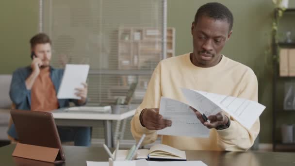 身穿浅黄色运动衫的黑人男子在工作场所分析财务文件的速度略为缓慢 而他的白人男性同事则在与背景客户的手机交谈中分析财务文件 — 图库视频影像