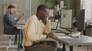 Gündelik kıyafet ticareti yapan Afrikalı Amerikalı erkek tüccar bilgisayar üzerinde online borsa analizi yapıyor ve müşterilerle telefonda konuşuyor, Kafkasyalı erkek meslektaşıyla birlikte çalışıyor.