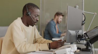 Gündelik giyim ve kulaklık takan Afro-Amerikan borsa simsarı bilgisayar monitörünün önünde oturmuş online borsayı analiz ediyor ve müşterilerle işbirliği yapıyor.