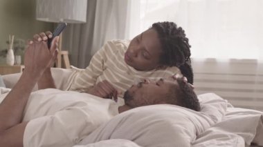 Afrika kökenli, aşık genç bir çift hafta sonu yatakta uzanıp akıllı telefonlara bakıyorlar.