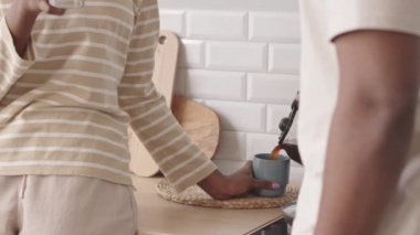 Sabahları kahvesini bardağa dolduran ve kız arkadaşıyla konuşan, minimalist mutfakta yan yana duran siyahi bir adam.