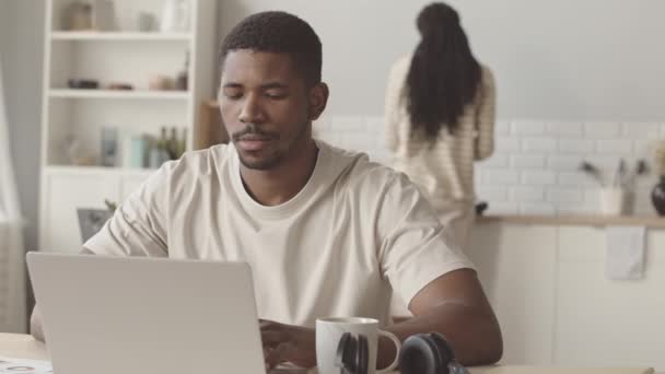 在他的女朋友为他做早餐的背景下 坐在厨房桌子旁 坐在笔记本电脑上工作的专注的黑人年轻人的慢动作 — 图库视频影像