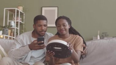 Genç Afrikalı Amerikalı çiftin yavaş yavaş robotik elektrik süpürgesini akıllı telefona bağlarken aynı anda oturma odasındaki kanepede oturup birlikte ev işi yapıyorlar.