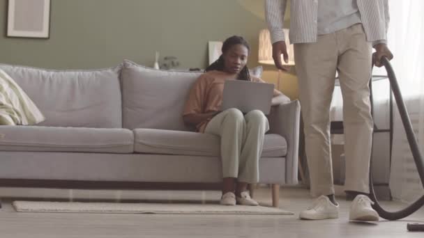 年轻的黑人男子在客厅里吸尘地板 而他的女朋友坐在沙发上使用笔记本电脑时 他的动作慢些 — 图库视频影像