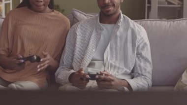 Genç ve neşeli Afrikalı Amerikalı çiftin video oyunları oynarken kontrolörleri kullanması, minimalist eko tarzı oturma odasında koltukta oturması.