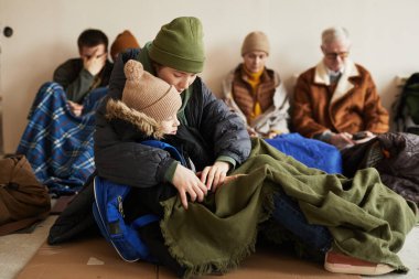 Mülteci barınağında saklanırken ve yerde battaniyelerle otururken uyuyan oğlunu tutan genç annenin yan görüntüsü.