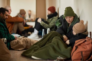 Mülteci barınağında çocuğu olan beyaz bir anne yerde oturuyor ve battaniyelerle kaplanmış ısınmaya çalışıyor.