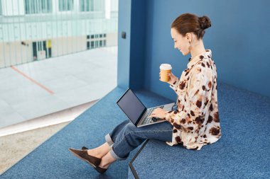 Mavi ofis alanında çalışırken dizüstü bilgisayar kullanan ve kahvenin tadını çıkaran genç bir profesyonelin grafiksel portresi.