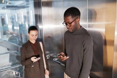 Modern ofis binasının asansöründe iş arkadaşıyla birlikte akıllı telefon kullanan siyahi iş adamının yan görünümü.