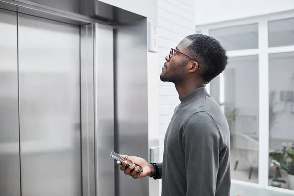 Minimal side view of young black businessman waiting for elevator in office building and holding phone, copy space