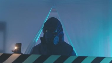 Koruyucu tulumlar, gözlükler ve solunum maskesi takan erkek biyoteknoloji müfettişinin akıllı telefonuna meşale tutarak acil servisteki gizli laboratuvarda polietilen kullanarak orta derecede yavaşlaması.