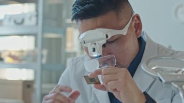 Modern laboratuarda bilgisayar çipini inceleyen gözlerinde lens ve beyaz önlük olan Asyalı erkek mühendisin orta boy yakın çekimi.