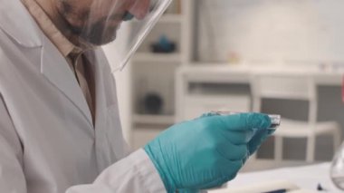Çalışma kıyafetleri içindeki Kafkas erkek bilim adamının yavaş yavaş petri kabındaki mavi sıvı maddeyi incelemesini, pipetle cam şişeye atmasını, laboratuvarda asistanla çalışmasını.