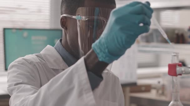 穿着防护面罩 实验室外套和手套的年轻黑人男性化学家在用某种液态物质加热蒸馏瓶中的实验研究 — 图库视频影像