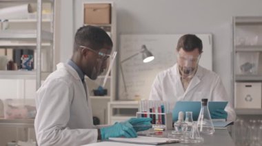 Laboratuvar önlükleri ve koruyucu yüz kalkanları takan iki farklı erkek kimyagerin orta boy yavaşlığı üzerinde cam ekipmanlarla masada oturması ve modern laboratuardaki maddelerle araştırma yapması.